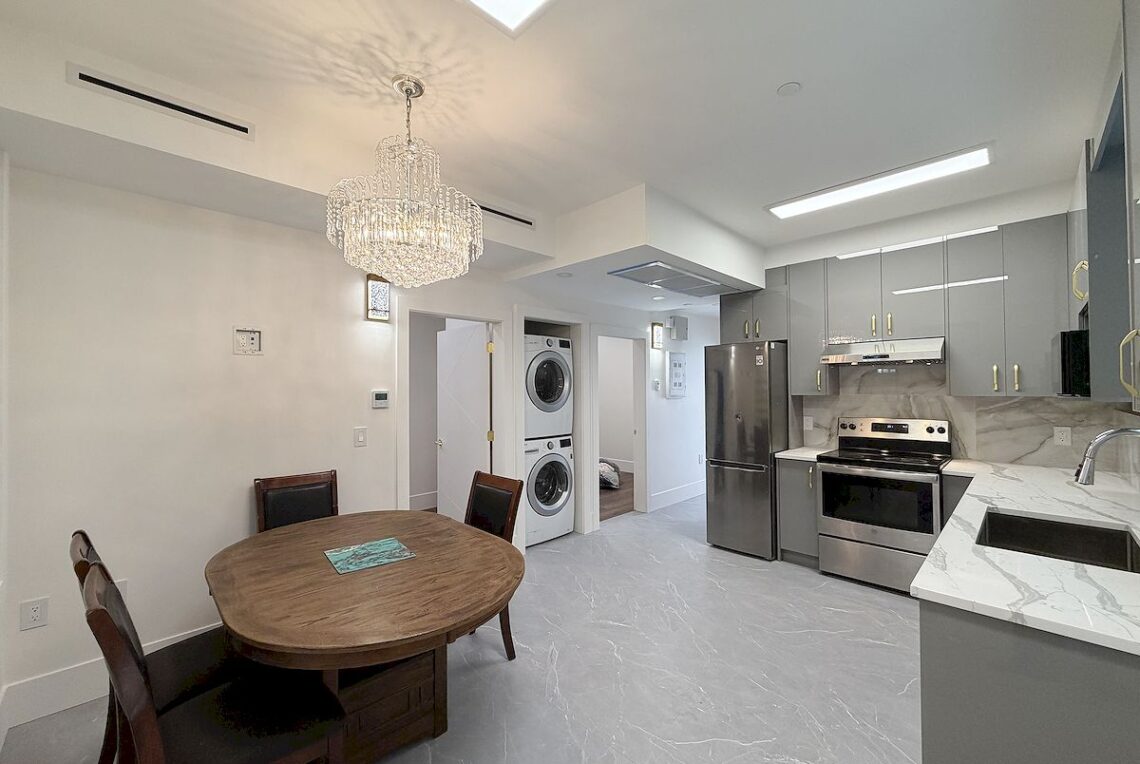 Ground Floor 3-Bed Suite in Vancouver East (Near Moberly Park) 溫東平地3房套房近 Moberly 公園 | Selling & Managing Vancouver Real Estate Modern kitchen and dining area with gray cabinets, stainless steel fridge and range, marble countertops, and a wooden round dining table under a crystal chandelier.