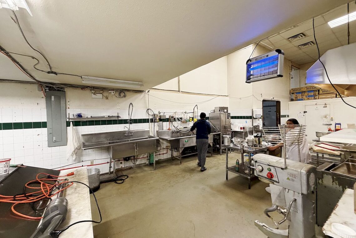 Vancouver 5,008 Sqft Food Processing Plant in Marpole For Sale 溫西Marpole 稀缺5,008平方英尺食品工場出售 | Selling & Managing Vancouver Real Estate Commercial kitchen with stainless steel sinks, prep tables, and a stand mixer; two workers at stations.
