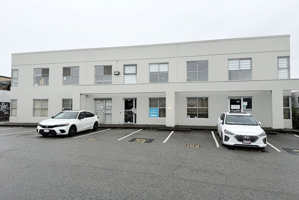 Rare 5,008 Sqft Industrial Corner Unit with Commercial Kitchen in Marpole 溫哥華 Marpole 稀缺5,008平方英尺工業轉角單元配備商業廚房 | Selling & Managing Vancouver Real Estate Two white cars parked in front of a gray office building with a blue entrance sign and multiple windows on both floors.