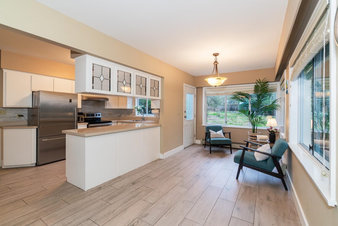 Vancouver West: 4 Bed 4 Bath House Near Magee School 溫西4睡房4衛浴獨立屋近Magee中學 | Selling & Managing Vancouver Real Estate Open kitchen and living area with white cabinetry, stainless steel appliances, a beige wall, and a breakfast island; teal chairs nearby by large windows.
