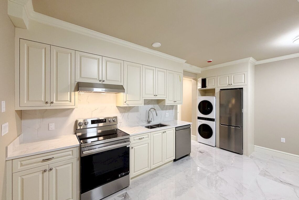 Brand New 2-Bed Suite in Vancouver West (Near Dunbar C.C.) 溫西全新兩房近Dunbar 社區中心 | Selling & Managing Vancouver Real Estate White kitchen with tall cabinets, stainless steel stove and range hood, marble countertops, and a laundry area with stacked washer and dryer beside a refrigerator.
