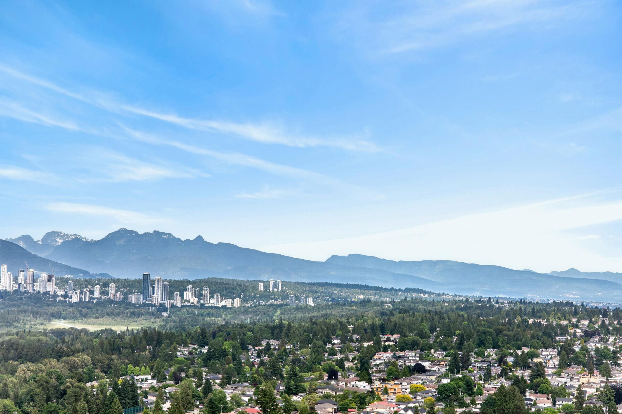 Metro Town Mountain View 2-Bed Condo Near Skytrain Station 鐵道鎮高層山景兩房公寓近天車站 | Selling & Managing Vancouver Real Estate City skyline at the foot of a snow-dusted mountain range under a clear blue sky, with dense trees and residential neighborhoods in the foreground.