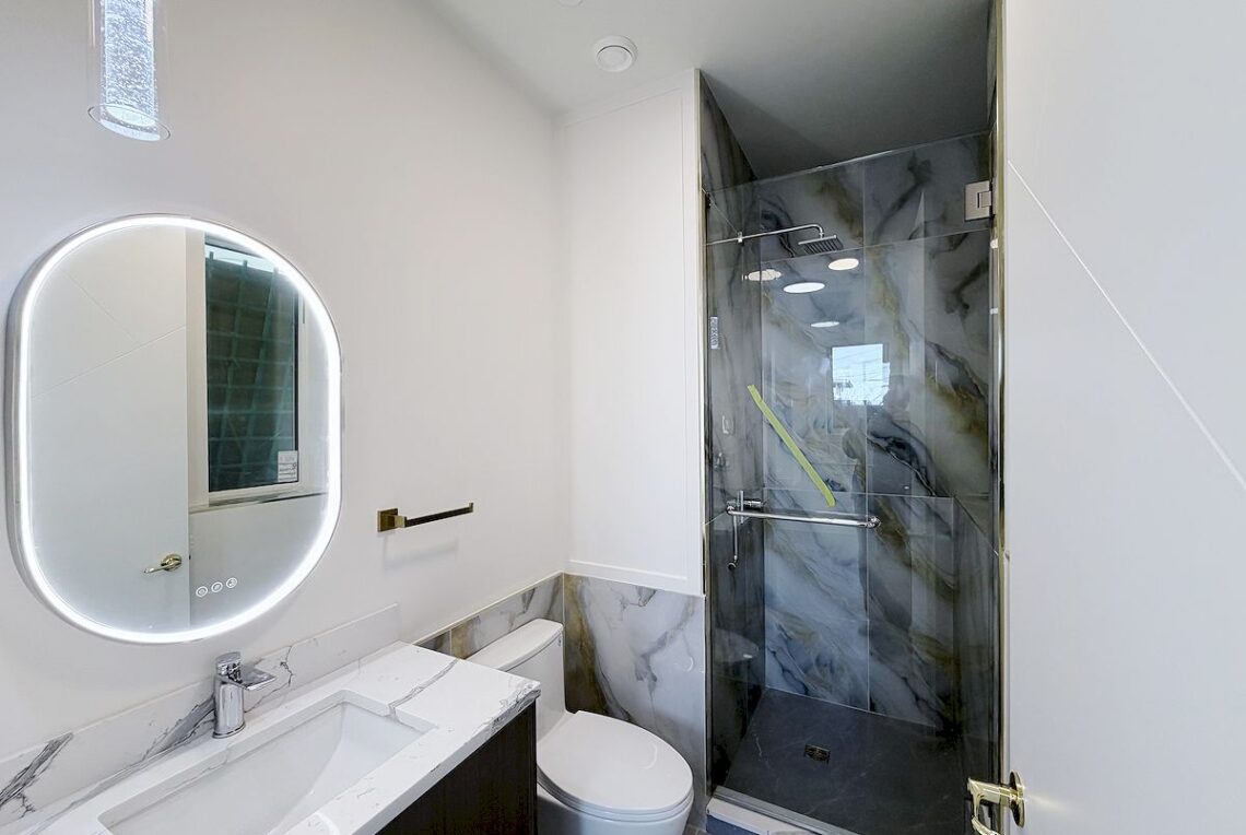 Ground Floor 1-Bed Suite in Vancouver East (Near Moberly Park) 溫東平地一房套房近 Moberly 公園 | Selling & Managing Vancouver Real Estate Round backlit mirror above marble countertop sink in a modern bathroom with a white wall and dark wood vanity.