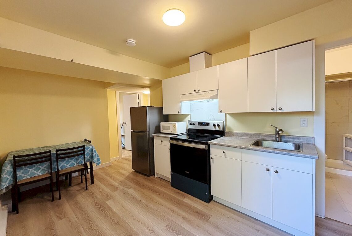 Richmond 3 Bed House Near McMath School 列治文McMath校區两房套房出租 | Selling & Managing Vancouver Real Estate Kitchen with white cabinets, stainless fridge, microwave, stove, sink, and a small two-chair dining area on the left with a blue tablecloth.