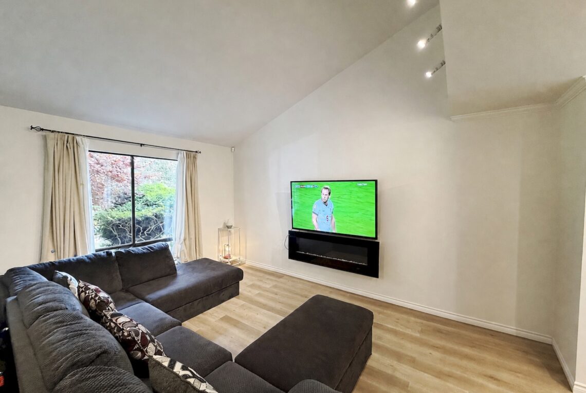 Richmond Furnished 4 Bed House Near Diefenbaker School 列治文帶家具四房獨立屋近Diefenbaker 小學 | Selling & Managing Vancouver Real Estate Modern living room with a dark gray sectional sofa near a window with cream curtains and a view outside. A wall-mounted TV sits above a fireplace shelf.