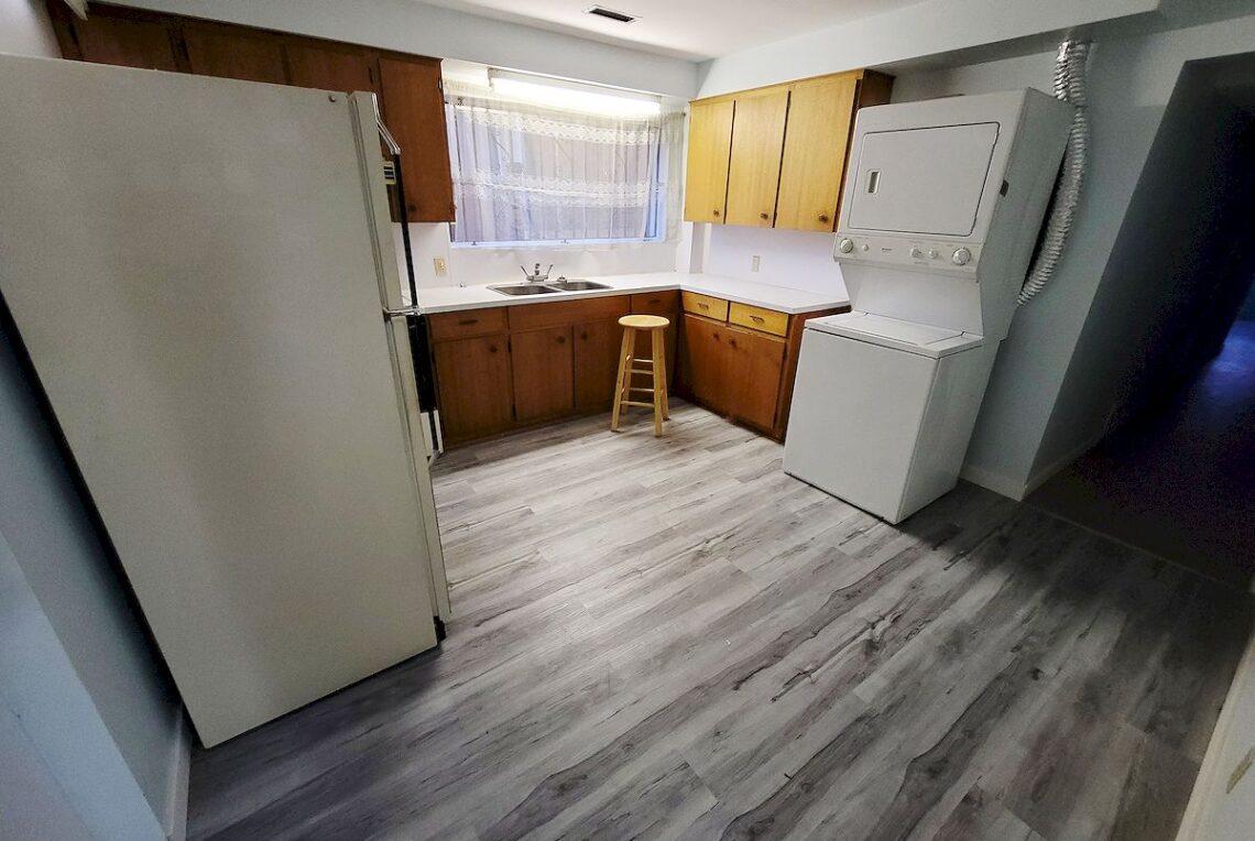 Vancouver 3-Bed Ground Floor Suite Near Joyce Skytrain 出租溫東獨立屋平地3房(近Joyce 天車) | Selling & Managing Vancouver Real Estate Basement kitchenette with white fridge on the left, stacked washer/dryer on the right, wooden cabinets, a sink under a lace-curtained window, and a small wooden stool.