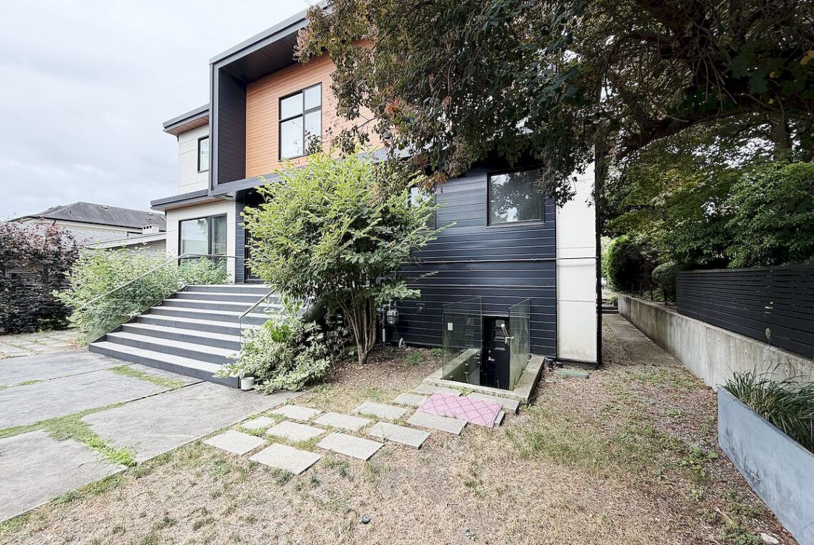 Vancouver 2-Bed Basement Suite Near Churchill School 溫西獨立屋土庫2房(近邱吉爾中學)出租 | Selling & Managing Vancouver Real Estate Modern two-story house with dark siding, a wooden accent panel, and a wide front staircase leading to the entry amidst greenery.