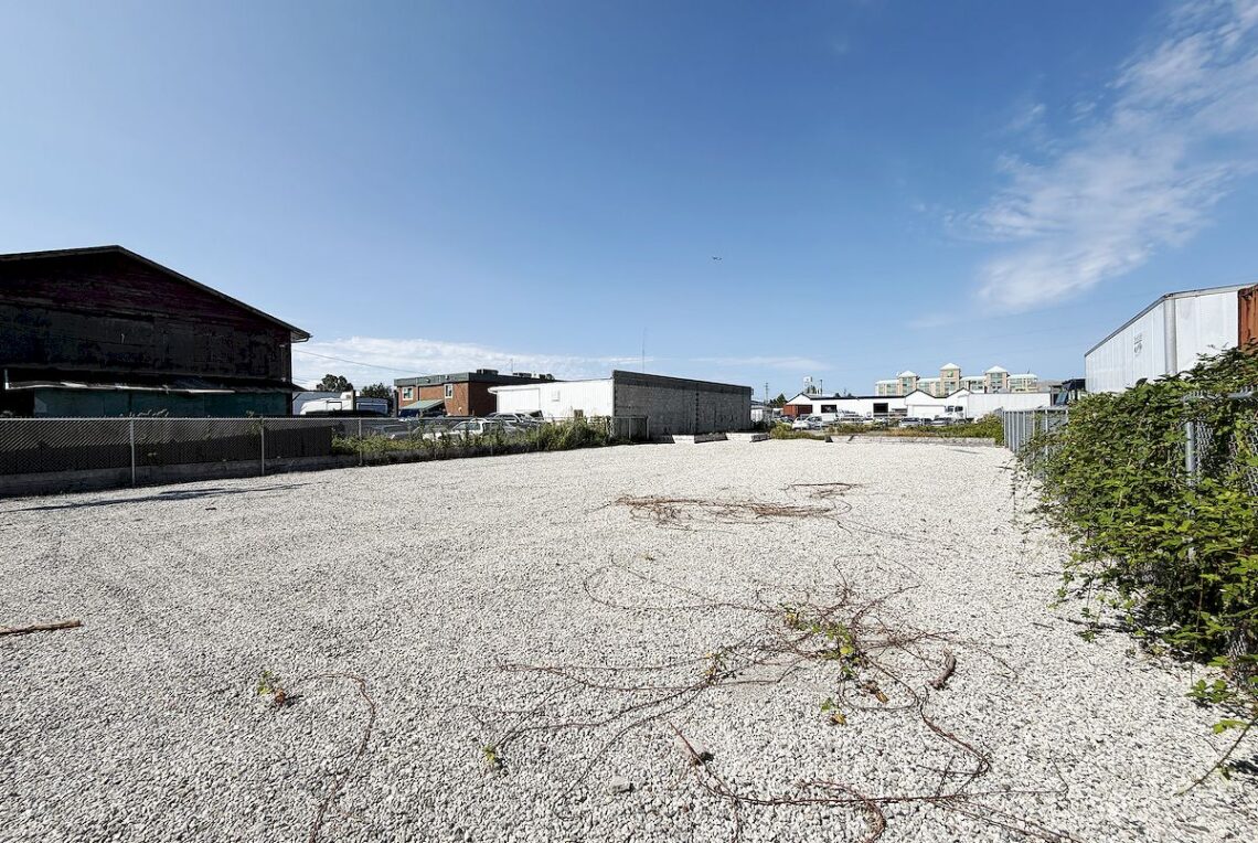 For Lease: Industrial land in Richmond (East Cambie) 列治文東甘比區工業地招租 | Selling & Managing Vancouver Real Estate Wide view of an empty gravel lot with sparse vegetation and tangled vines, industrial buildings in the background under a blue sky.